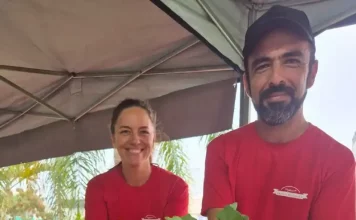 Casal de agricultores usando camisetas vermelhas atende clientes atrás do balcão da feira orgânica em São José. Na frente, há uma variedade de produtos frescos e naturais, como alface, repolho roxo, repolho verde, cenoura, batata-doce, rabanete e banana, destacando a diversidade da agricultura familiar local.