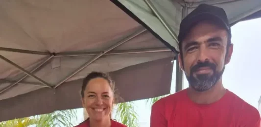 Casal de agricultores usando camisetas vermelhas atende clientes atrás do balcão da feira orgânica em São José. Na frente, há uma variedade de produtos frescos e naturais, como alface, repolho roxo, repolho verde, cenoura, batata-doce, rabanete e banana, destacando a diversidade da agricultura familiar local.