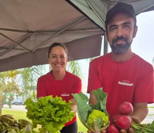Casal de agricultores usando camisetas vermelhas atende clientes atrás do balcão da feira orgânica em São José. Na frente, há uma variedade de produtos frescos e naturais, como alface, repolho roxo, repolho verde, cenoura, batata-doce, rabanete e banana, destacando a diversidade da agricultura familiar local.