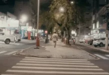 Pessoa caminha à noite pelo calçadão iluminado do centro de São José, SC, cenário do filme Peça para Solidão Solo.