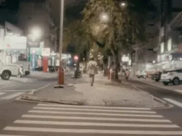 Pessoa caminha à noite pelo calçadão iluminado do centro de São José, SC, cenário do filme Peça para Solidão Solo.