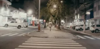 Pessoa caminha à noite pelo calçadão iluminado do centro de São José, SC, cenário do filme Peça para Solidão Solo.