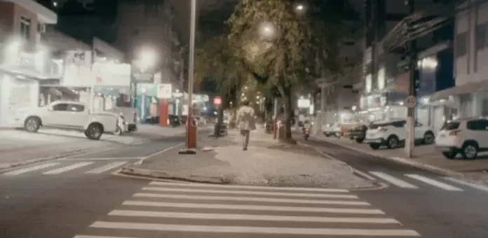 Pessoa caminha à noite pelo calçadão iluminado do centro de São José, SC, cenário do filme Peça para Solidão Solo.