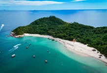 Foto aérea mostra a Ilha do Campeche cercada pelo mar azul. Pequenos barcos estão próximos à costa, enquanto pessoas aproveitam o dia na areia clara da praia. A vegetação verde cobre boa parte da ilha, destacando sua beleza natural.