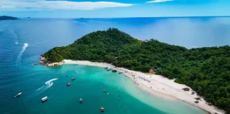 Foto aérea mostra a Ilha do Campeche cercada pelo mar azul. Pequenos barcos estão próximos à costa, enquanto pessoas aproveitam o dia na areia clara da praia. A vegetação verde cobre boa parte da ilha, destacando sua beleza natural.
