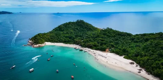 Foto aérea mostra a Ilha do Campeche cercada pelo mar azul. Pequenos barcos estão próximos à costa, enquanto pessoas aproveitam o dia na areia clara da praia. A vegetação verde cobre boa parte da ilha, destacando sua beleza natural.