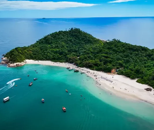 Foto aérea mostra a Ilha do Campeche cercada pelo mar azul. Pequenos barcos estão próximos à costa, enquanto pessoas aproveitam o dia na areia clara da praia. A vegetação verde cobre boa parte da ilha, destacando sua beleza natural.