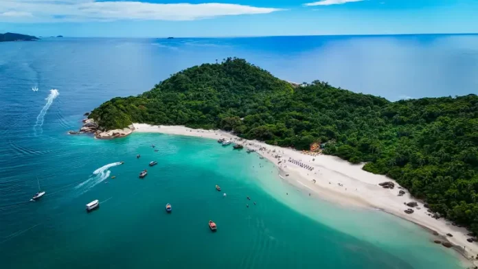 Foto aérea mostra a Ilha do Campeche cercada pelo mar azul. Pequenos barcos estão próximos à costa, enquanto pessoas aproveitam o dia na areia clara da praia. A vegetação verde cobre boa parte da ilha, destacando sua beleza natural.