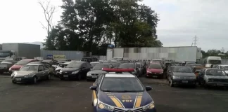 A imagem mostra, em primeiro plano, uma viatura azul da Polícia Rodoviária Federal (PRF) com faixas amarelas nas laterais. No capô, destaca-se o símbolo oficial da PRF. Ao fundo, vários carros apreendidos estão alinhados e estacionados lado a lado em um pátio aberto. O ambiente é externo, sob luz natural, e o pátio possui piso de concreto.