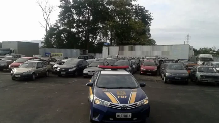 A imagem mostra, em primeiro plano, uma viatura azul da Polícia Rodoviária Federal (PRF) com faixas amarelas nas laterais. No capô, destaca-se o símbolo oficial da PRF. Ao fundo, vários carros apreendidos estão alinhados e estacionados lado a lado em um pátio aberto. O ambiente é externo, sob luz natural, e o pátio possui piso de concreto.