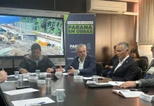 Três secretários de estado estão sentados ao redor de uma mesa de reuniões. Sobre a mesa, há papéis, canetas e documentos. Ao fundo, um grande painel exibe a imagem de uma obra em andamento e traz os dizeres "Paraná em Obras".