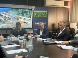 Três secretários de estado estão sentados ao redor de uma mesa de reuniões. Sobre a mesa, há papéis, canetas e documentos. Ao fundo, um grande painel exibe a imagem de uma obra em andamento e traz os dizeres "Paraná em Obras".