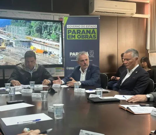 Três secretários de estado estão sentados ao redor de uma mesa de reuniões. Sobre a mesa, há papéis, canetas e documentos. Ao fundo, um grande painel exibe a imagem de uma obra em andamento e traz os dizeres "Paraná em Obras".
