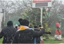 Pessoas vestidas com casacos grossos e roupas de inverno observam a neve caindo. Elas estão de pé, voltadas para a paisagem, enquanto os flocos descem suavemente do céu. O clima transmite frio e encantamento