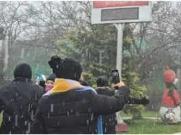 Pessoas vestidas com casacos grossos e roupas de inverno observam a neve caindo. Elas estão de pé, voltadas para a paisagem, enquanto os flocos descem suavemente do céu. O clima transmite frio e encantamento