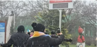 Pessoas vestidas com casacos grossos e roupas de inverno observam a neve caindo. Elas estão de pé, voltadas para a paisagem, enquanto os flocos descem suavemente do céu. O clima transmite frio e encantamento