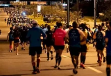 Pessoas correm à noite pela Avenida Beira-Mar Continental, em Florianópolis, durante a Meia Maratona Noturna. As ruas estão iluminadas, e os corredores usam roupas esportivas com números de inscrição.