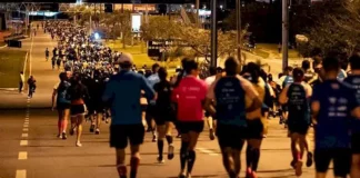 Pessoas correm à noite pela Avenida Beira-Mar Continental, em Florianópolis, durante a Meia Maratona Noturna. As ruas estão iluminadas, e os corredores usam roupas esportivas com números de inscrição.