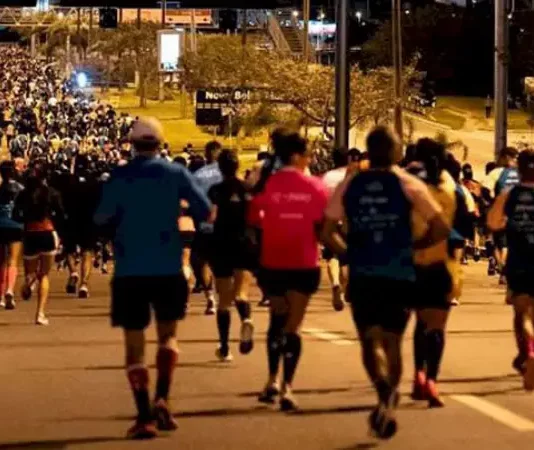 Pessoas correm à noite pela Avenida Beira-Mar Continental, em Florianópolis, durante a Meia Maratona Noturna. As ruas estão iluminadas, e os corredores usam roupas esportivas com números de inscrição.