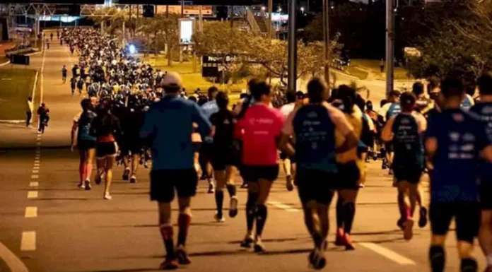 Pessoas correm à noite pela Avenida Beira-Mar Continental, em Florianópolis, durante a Meia Maratona Noturna. As ruas estão iluminadas, e os corredores usam roupas esportivas com números de inscrição.