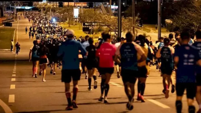 Pessoas correm à noite pela Avenida Beira-Mar Continental, em Florianópolis, durante a Meia Maratona Noturna. As ruas estão iluminadas, e os corredores usam roupas esportivas com números de inscrição.