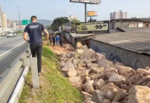 Homem de camiseta azul caminhando na marginal da Via Expressa, ao lado de pedras pontiagudas colocadas pela prefeitura para evitar ocupações irregulares.