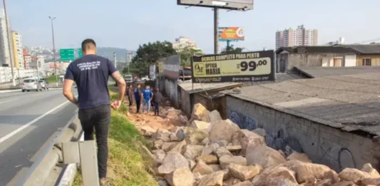 Homem de camiseta azul caminhando na marginal da Via Expressa, ao lado de pedras pontiagudas colocadas pela prefeitura para evitar ocupações irregulares.