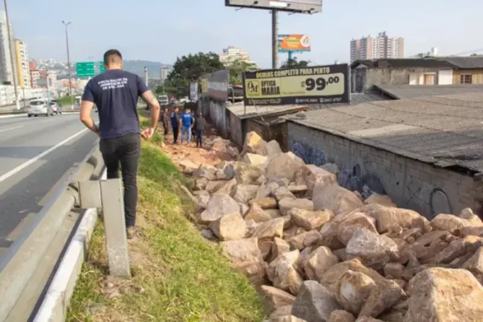 Homem de camiseta azul caminhando na marginal da Via Expressa, ao lado de pedras pontiagudas colocadas pela prefeitura para evitar ocupações irregulares.