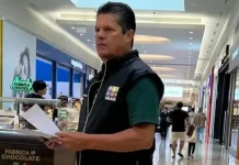 O diretor do Procon de São José, Tetê Souza, está de pé no corredor de um shopping. Ele veste uma camiseta verde e um colete preto com o logotipo do Procon. Tetê segura algumas folhas de papel nas mãos e está em frente a um quiosque, em atitude atenta, como se realizasse uma fiscalização.