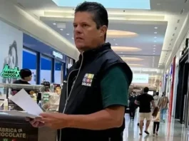 O diretor do Procon de São José, Tetê Souza, está de pé no corredor de um shopping. Ele veste uma camiseta verde e um colete preto com o logotipo do Procon. Tetê segura algumas folhas de papel nas mãos e está em frente a um quiosque, em atitude atenta, como se realizasse uma fiscalização.