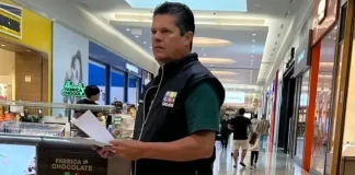 O diretor do Procon de São José, Tetê Souza, está de pé no corredor de um shopping. Ele veste uma camiseta verde e um colete preto com o logotipo do Procon. Tetê segura algumas folhas de papel nas mãos e está em frente a um quiosque, em atitude atenta, como se realizasse uma fiscalização.