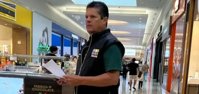 O diretor do Procon de São José, Tetê Souza, está de pé no corredor de um shopping. Ele veste uma camiseta verde e um colete preto com o logotipo do Procon. Tetê segura algumas folhas de papel nas mãos e está em frente a um quiosque, em atitude atenta, como se realizasse uma fiscalização.