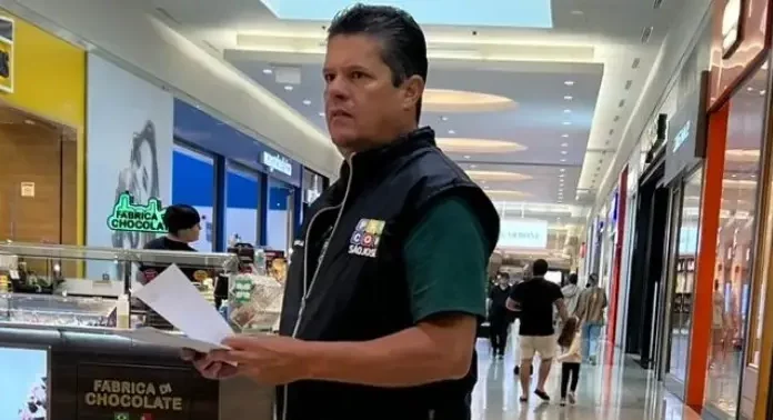 O diretor do Procon de São José, Tetê Souza, está de pé no corredor de um shopping. Ele veste uma camiseta verde e um colete preto com o logotipo do Procon. Tetê segura algumas folhas de papel nas mãos e está em frente a um quiosque, em atitude atenta, como se realizasse uma fiscalização.