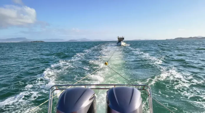 Vista da popa (parte traseira) da lancha dos bombeiros em alto mar, rebocando uma embarcação pesqueira à deriva. O mar está calmo, e o céu claro