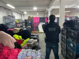 Polícia Civil apreende R$ 20 milhões em roupas falsificadas em operação em SC Foto mostra um policial civil visto de costas. Ele veste uma jaqueta preta e, por cima dela, um colete à prova de balas com a inscrição “DEIC Polícia Civil” nas costas. O policial usa calça jeans azul, carrega uma arma na cintura e está cercado por diversas peças de roupas falsificadas armazenadas dentro de um galpão.