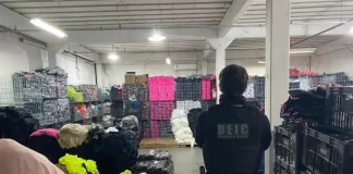 Foto mostra um policial civil visto de costas. Ele veste uma jaqueta preta e, por cima dela, um colete à prova de balas com a inscrição “DEIC Polícia Civil” nas costas. O policial usa calça jeans azul, carrega uma arma na cintura e está cercado por diversas peças de roupas falsificadas armazenadas dentro de um galpão.