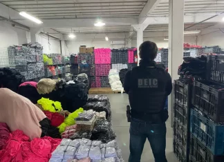 Foto mostra um policial civil visto de costas. Ele veste uma jaqueta preta e, por cima dela, um colete à prova de balas com a inscrição “DEIC Polícia Civil” nas costas. O policial usa calça jeans azul, carrega uma arma na cintura e está cercado por diversas peças de roupas falsificadas armazenadas dentro de um galpão.