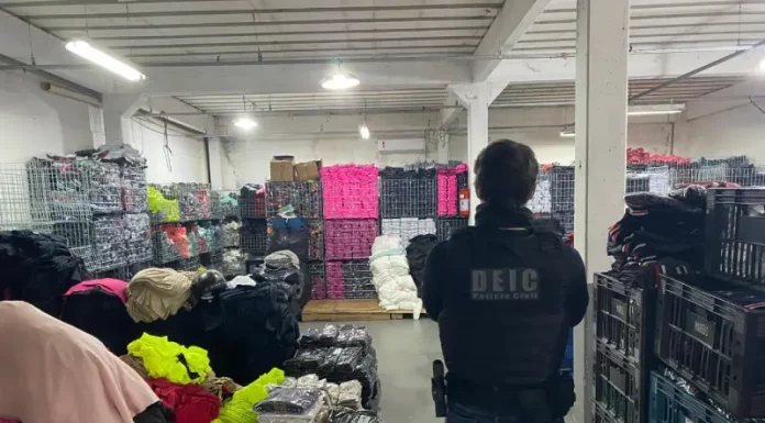 Foto mostra um policial civil visto de costas. Ele veste uma jaqueta preta e, por cima dela, um colete à prova de balas com a inscrição “DEIC Polícia Civil” nas costas. O policial usa calça jeans azul, carrega uma arma na cintura e está cercado por diversas peças de roupas falsificadas armazenadas dentro de um galpão.