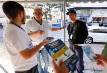 Homem de camiseta branca segurando um panfelto do Trato Pelo Araujo, conversanbdo com um senhor de camisa branca, usando óculos e com um homem de camisa preta e boné