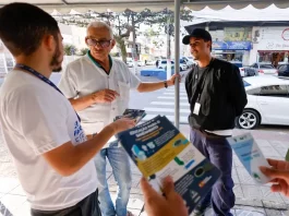 Homem de camiseta branca segurando um panfelto do Trato Pelo Araujo, conversanbdo com um senhor de camisa branca, usando óculos e com um homem de camisa preta e boné