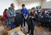 Homem vestindo jaqueta cinza com a logo do Governo do Estado segura uma jaqueta preta de uniforme escolar. Atrás dele, um menino com moletom azul observa. Ao lado, uma menina com moletom preto segura outra jaqueta preta. Eles estão no ginásio de esportes de uma escola, onde vários alunos estão sentados ao fundo.