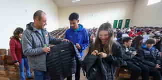 Homem vestindo jaqueta cinza com a logo do Governo do Estado segura uma jaqueta preta de uniforme escolar. Atrás dele, um menino com moletom azul observa. Ao lado, uma menina com moletom preto segura outra jaqueta preta. Eles estão no ginásio de esportes de uma escola, onde vários alunos estão sentados ao fundo.