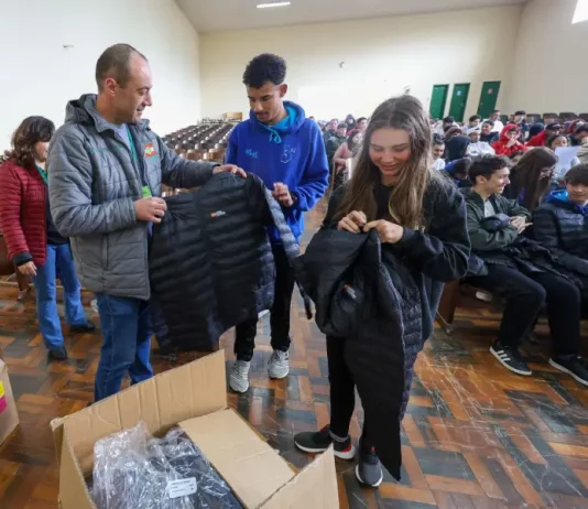 Homem vestindo jaqueta cinza com a logo do Governo do Estado segura uma jaqueta preta de uniforme escolar. Atrás dele, um menino com moletom azul observa. Ao lado, uma menina com moletom preto segura outra jaqueta preta. Eles estão no ginásio de esportes de uma escola, onde vários alunos estão sentados ao fundo.
