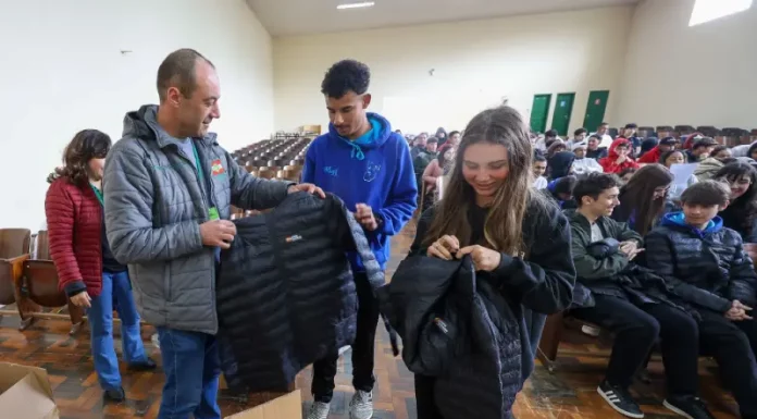 Homem vestindo jaqueta cinza com a logo do Governo do Estado segura uma jaqueta preta de uniforme escolar. Atrás dele, um menino com moletom azul observa. Ao lado, uma menina com moletom preto segura outra jaqueta preta. Eles estão no ginásio de esportes de uma escola, onde vários alunos estão sentados ao fundo.