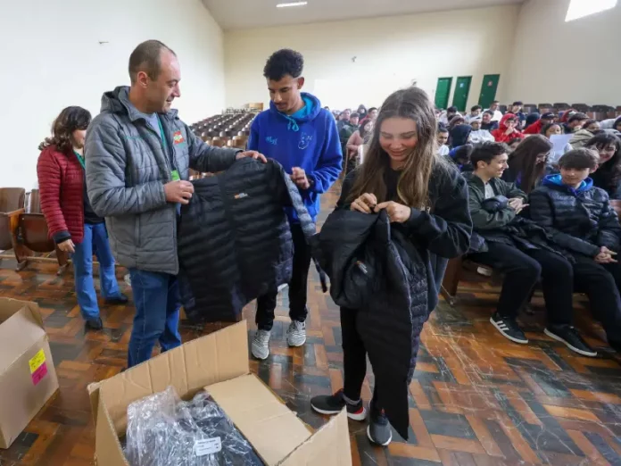 Homem vestindo jaqueta cinza com a logo do Governo do Estado segura uma jaqueta preta de uniforme escolar. Atrás dele, um menino com moletom azul observa. Ao lado, uma menina com moletom preto segura outra jaqueta preta. Eles estão no ginásio de esportes de uma escola, onde vários alunos estão sentados ao fundo.