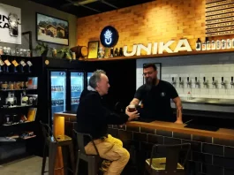 Homem aprecia cerveja artesanal no balcão do bar da Unika, em Coqueiros, enquanto atendente entrega o copo.