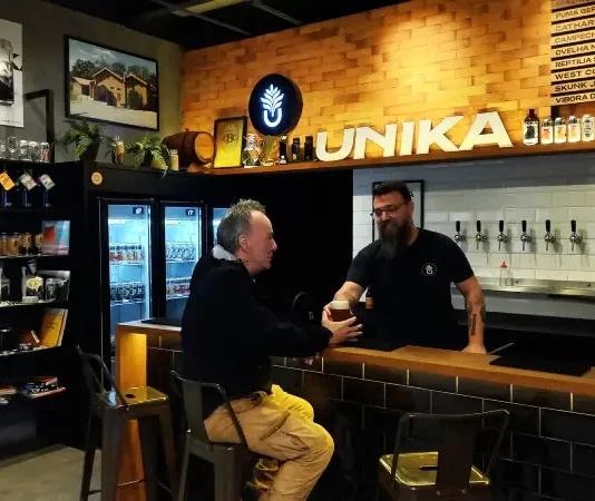 Homem aprecia cerveja artesanal no balcão do bar da Unika, em Coqueiros, enquanto atendente entrega o copo.