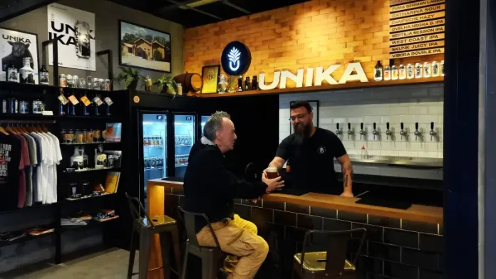 Homem aprecia cerveja artesanal no balcão do bar da Unika, em Coqueiros, enquanto atendente entrega o copo.