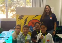 Professora posa com alunos em frente a uma maquete escolar, representando o processo seletivo emergencial na Educação de Florianópolis.