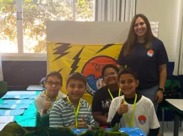 Professora posa com alunos em frente a uma maquete escolar, representando o processo seletivo emergencial na Educação de Florianópolis.